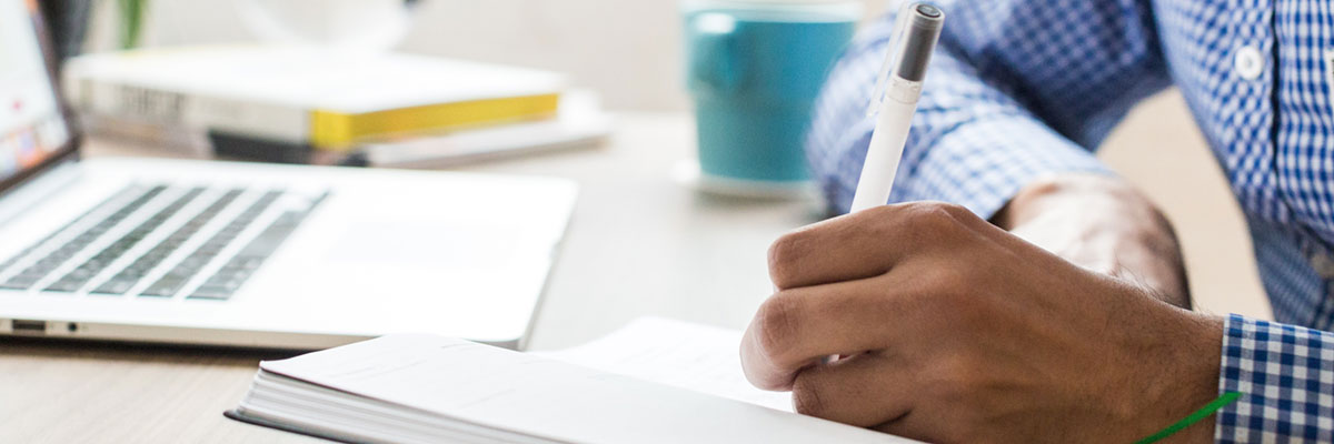 A man in a blue checkered shirt writing in a notebook in front of his laptop.